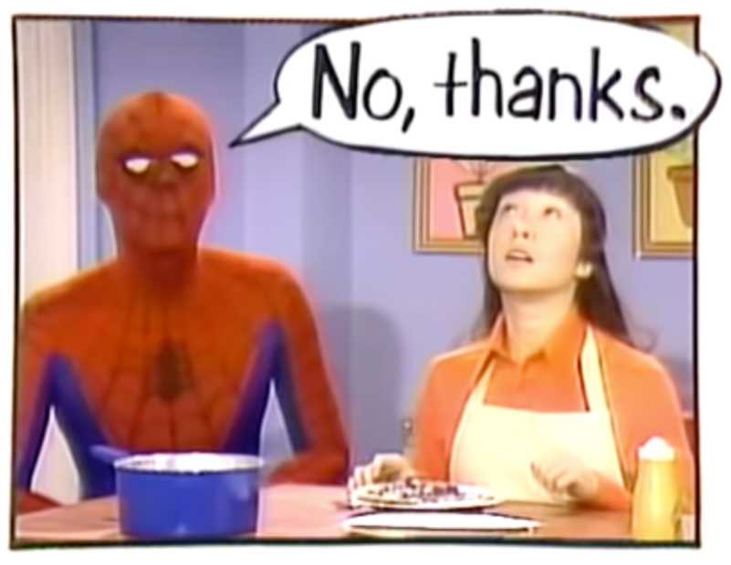 A shot from the Spider-Man segment of the Electric Company TV show. Spider-Man and a woman are in what looks like a kitchen. The woman is wearing an apron. The two are behind a tall table that has a pot and some plates with food on it. A large text bubble shows Spider-Man is saying “No, thanks.”